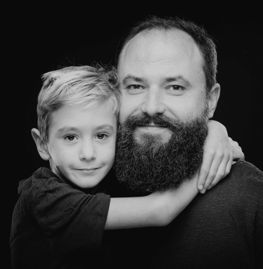 Herzliches Familienportrait vom Fotostudio Reuther, das einen Vater und seinen Sohn zeigt, die sich vor einem schwarzen Hintergrund umarmen.
