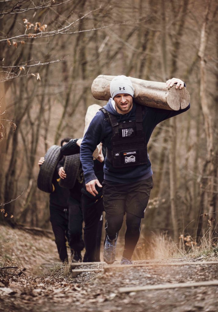 Männer, die durch den Wald klettern und dabei Baumstämme tragen, was ihre Kraft und Ausdauer in einer herausfordernden Outdoor-Workout-Situation zeigt.