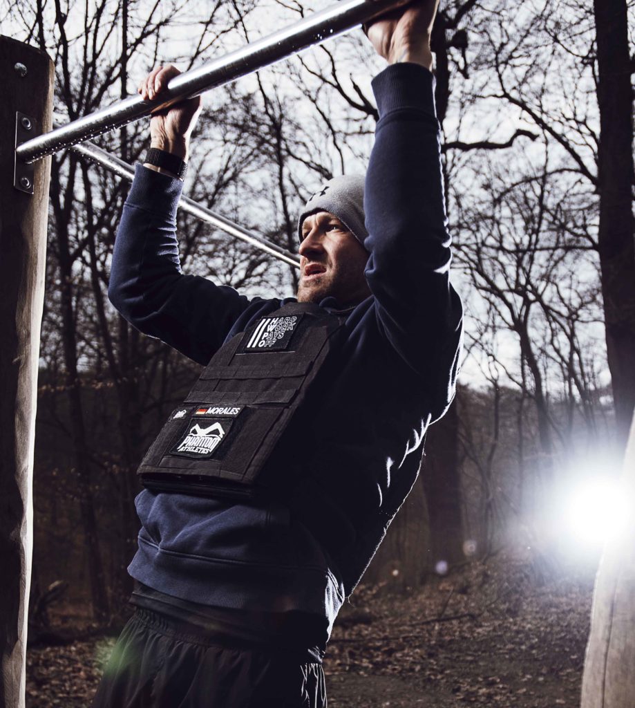 Ein Mann, der im Wald Klimmzüge an einem stabilen Baumstamm macht, während er ein Gewichtsgurt trägt, um seine Kraft und Fitness in einer natürlichen Umgebung zu steigern.