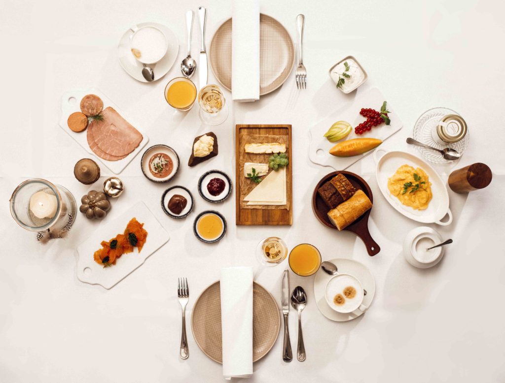 Symmetrische Detailaufnahme eines kompletten Frühstücks von oben, das verschiedene Speisen wie Brötchen, Obst, Joghurt und Getränke zeigt.