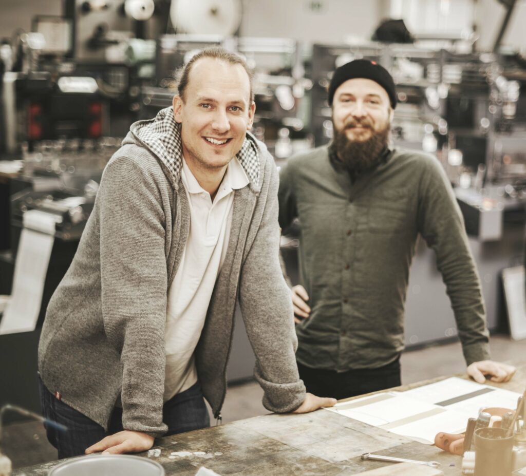 Portrait von Leiter und Mitarbeiter der Etikettierfirma vor dem Firmenhintergrund, lächelnd und professionell.