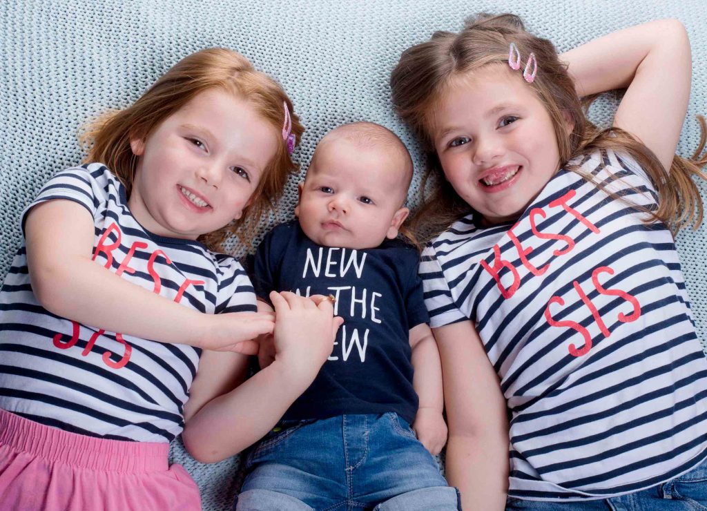 Fröhliches Familienportrait vom Fotostudio Reuther, das drei Geschwister zeigt, die auf dem Boden liegen und in die Kamera lachen.