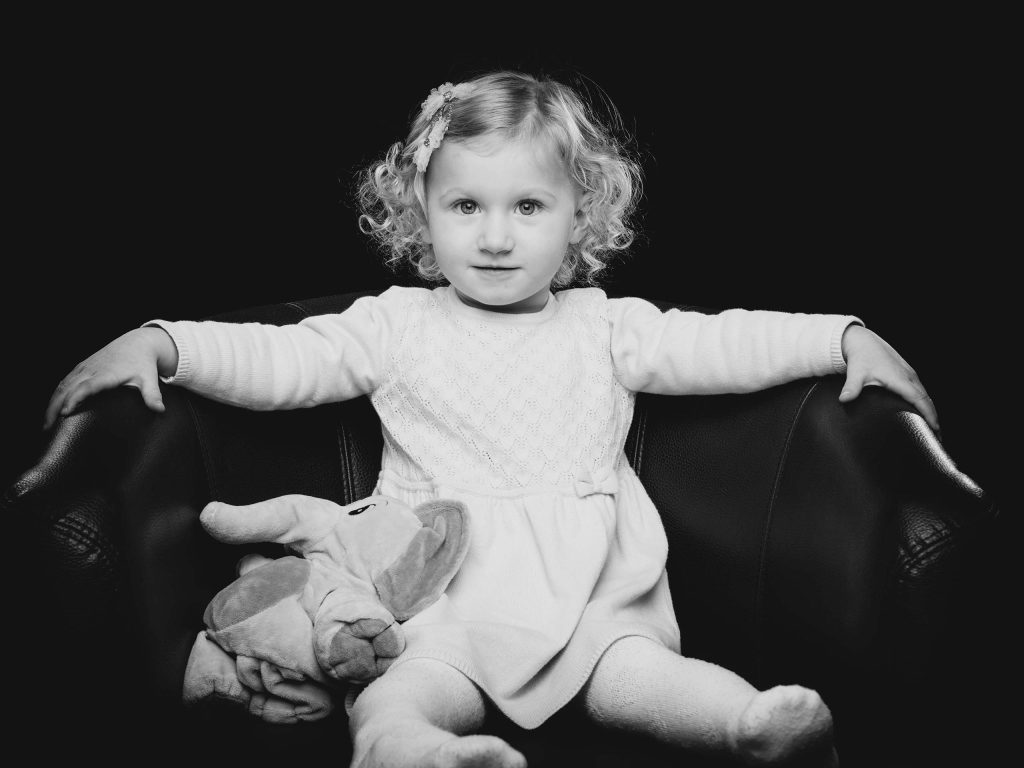 Charmantes Familienportrait vom Fotostudio Reuther, das ein kleines Mädchen zeigt, das selbstbewusst auf einem Sessel sitzt.