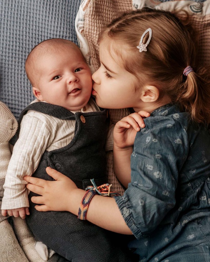 Süßes Familienportrait vom Fotostudio Reuther, das eine Schwester zeigt, die ihr Baby küsst, während das Baby in die Kamera lacht.