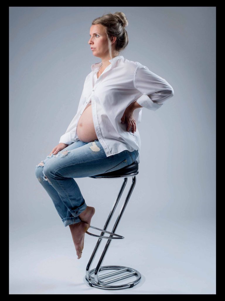 Studioaufnahme einer schwangeren Frau, die auf einem Hocker sitzt. Schwangerschaftsshooting bei Fotostudio Reuther.